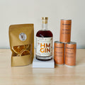 Bottle of HM Gin with a package of dehydrated orange wheels and cans of Sodasmith Australian Dry Ginger Ale on a wooden surface.