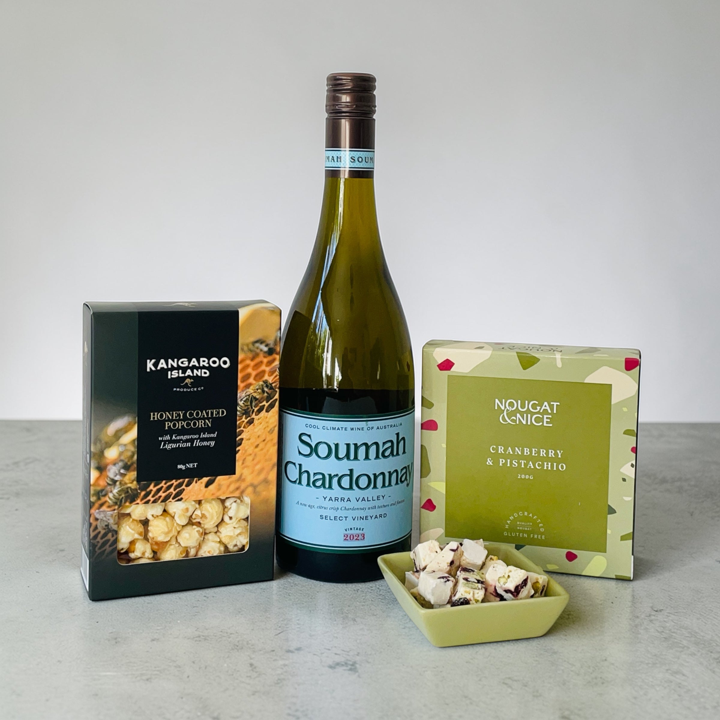 Bottle of Soumah Chardonnay with a box of kangaroo island honey coated popcorn and a box of nougat and nice cranberry and pistachio nougat on a gray surface.